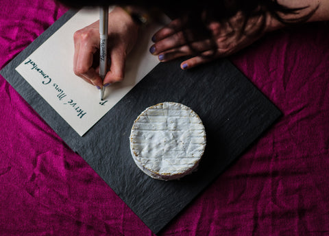 overhead shot of french camembert cheese on black board with hand writing cheese name on paper