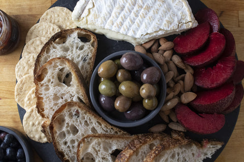 overhead view of round slate serving board with bread, bloomy cheese, plums, and olives