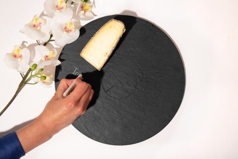 round slate serving board with cheese, flowers, and hand writing on the board with soapstone chalk