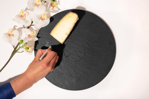 round slate serving board with cheese, flowers, and hand labeling it with soapstone chalk