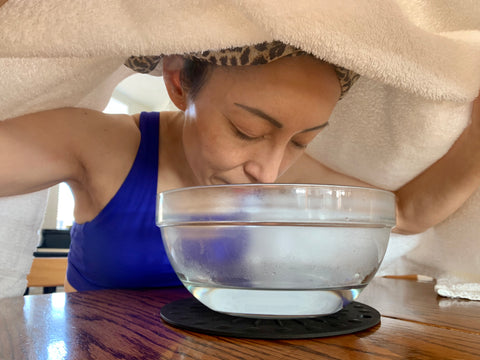 A woman steaming her face with bowl of water in front of her and a towel over her head
