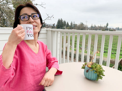 Woman with dark hair, in pink blouse sits at a table on a balcony, and holds a cup