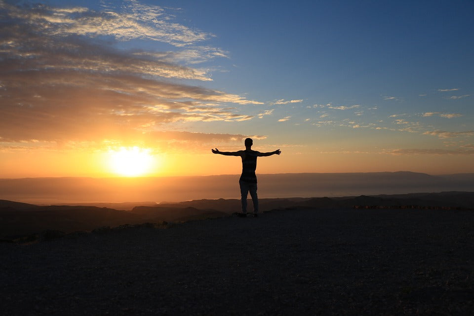 A man in silhouette stands looking towards the sunset with his arms outstretched positively.