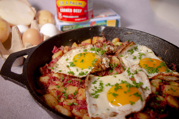 Corned beef hash with three fried sunny side up eggs garnished with parsely in a black cast iron skillet pan. In the background there is a carton of eggs with potatoes and a cut white onion next to a can of Ox and Palm corned beef and a box of butter.