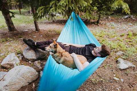 Man sitting in a blue hammock outside with his dog.