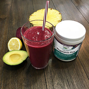 A cup of the detoxing Magnesium Smoothie next to an avocado, lemon, pineapple, and a tub of Ionic-Fizz Magnesium Plus