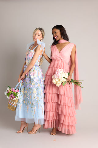 Duo shot of a soft pink maxi dress with self-tie bow ribbons as straps and a tiered tulle maxi skirt. Paired next to a soft blue midi dress with self-tie bow straps and 3D flowers along the bust and skirt of the flowy dress.