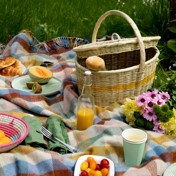 La Basketry handwoven picnic basket in yellow slightly open with a baguette inside surrounded by fruit and fresh flowers