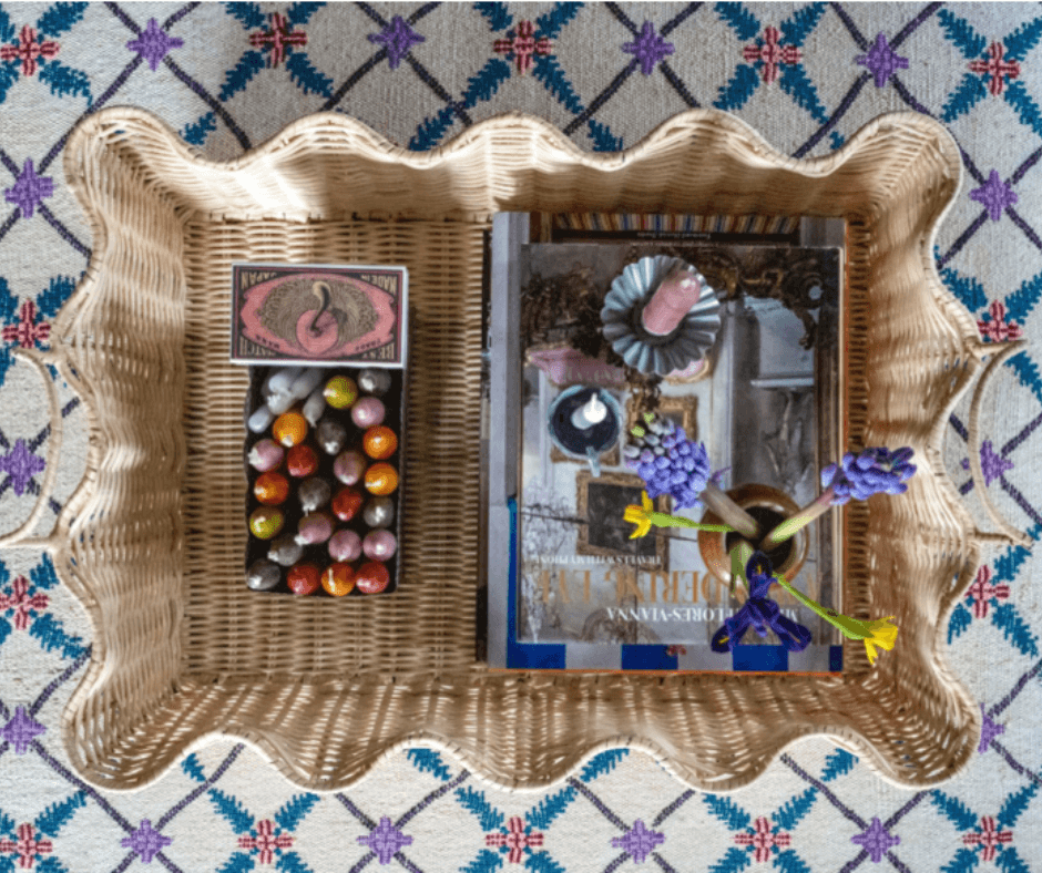 Top down view of a rattan ripple tray filled with candles, a magazine and flowers against a vintage patterned carpet, by Lisa Mehydene, founder of edit58. Image for her interview with La Basketry