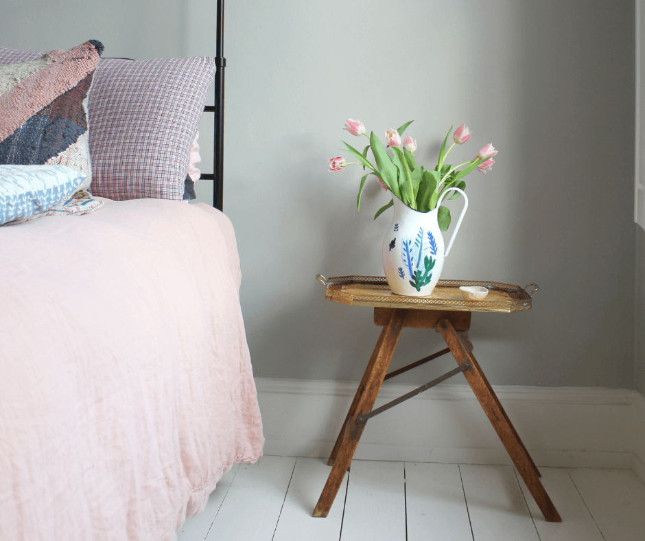 Close up of a bedside wooden table next to a bed with baby pink bedding, with a white vase decorated with blue flowers filled with pink tulips, from Lisa Mehydene, founder of edit58. Image for her interview with La Basketry