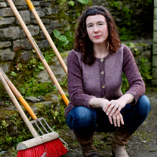woman crouching in front of garden tools with hands clasped in front wearing a pink and purple cardigan