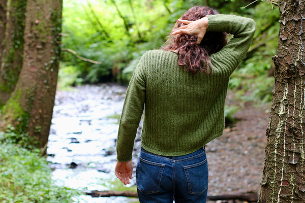 woman from back in woods, wearing a green sweater and lifting her hair up