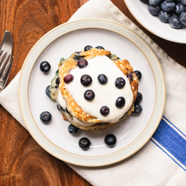 Blueberry pancakes stack on a white plate with a bowl of blueberries on a wooden table