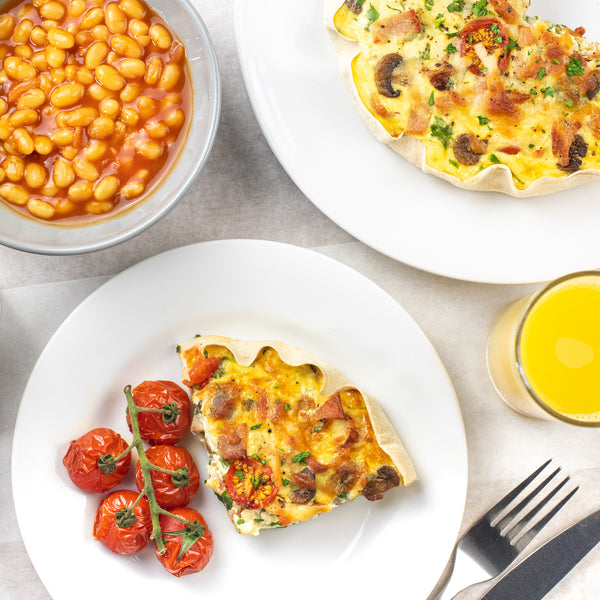 Breakfast quiche slice on a white plate with a bowl of beans and a glass of orange juice