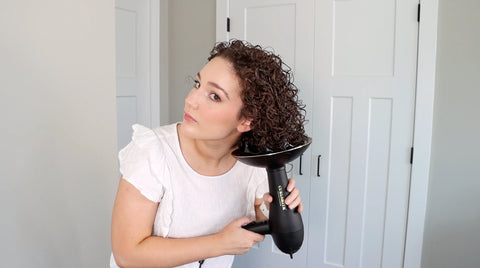 a curly haired woman in a white top holds her brown curls into the bowl of a diffuse