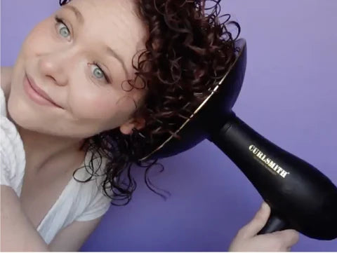 a curly haired woman holds her hair into a diffuser hair dryer in front of a purple backdrop