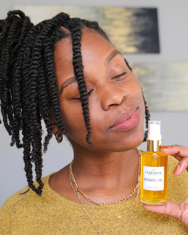 a woman with her type 4 hair in twists smiles with her eyes closed holding a bottle of hair oil
