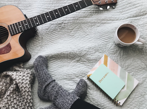 a person in cosy socks sitting on their bed with a journal, coffee, and guitar