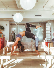 Dark haired woman does a yoga backbend in a sports bra and leggings in a sun lit room on top of a low table