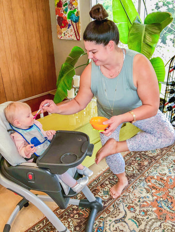 woman feeds child in high chair while doing standing figure 4