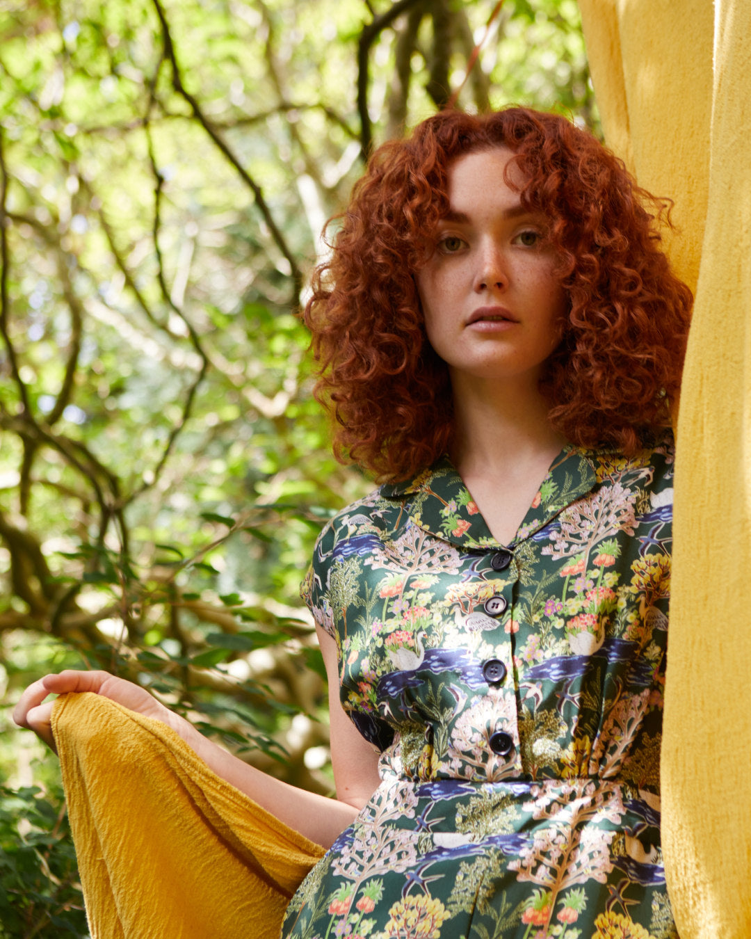 Ginger haired woman wearing a patterned, floral jumpsuit in a garden.