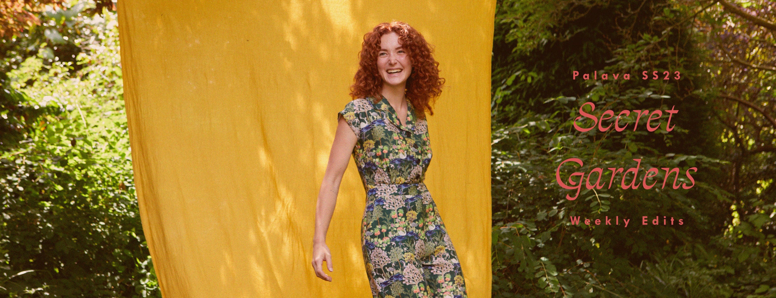 Woman smiling in a garden. She's wearing a dark green jumpsuit with a floral print that depicts botanical gardens.