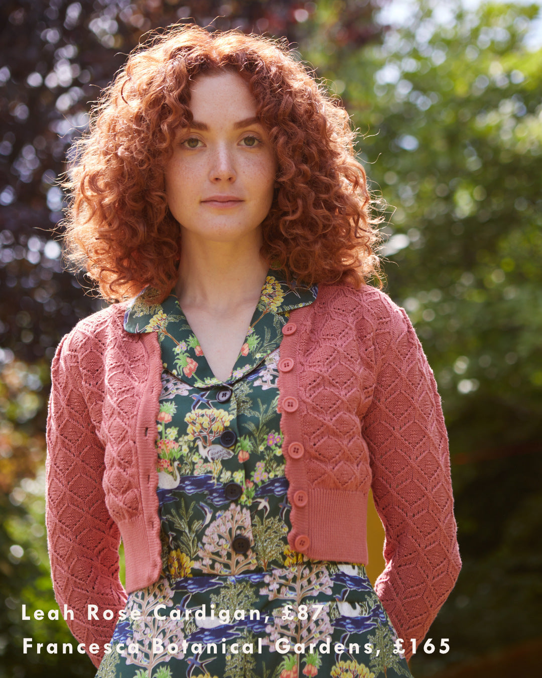 Ginger haired woman wearing a patterned, floral jumpsuit and a pink textured cardigan in a garden.