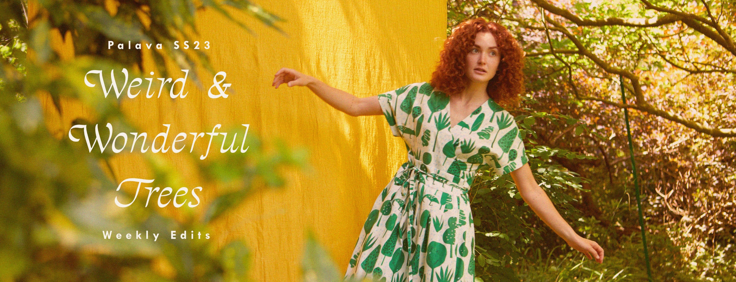 Woman with curly ginger hair with her arms stretched out. She's wearing an ivory wrap dress that has a green print depicting topiary trees.