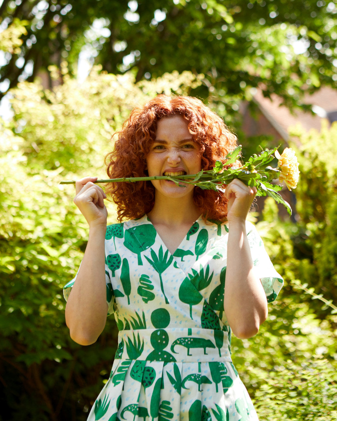 Ginger haired woman green and ivory printed wrap dress. She's biting on the step of a yellow flower.