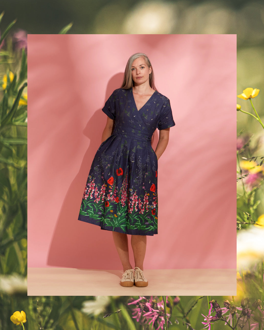 Woman wearing a printed wrap dress. The dress' border print of a dress. The border print depicts foxgloves, poppies and other flowers on an indigo blue background.