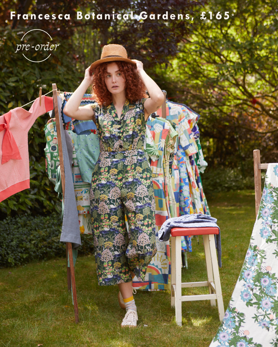 Ginger haired woman wearing a patterned, floral jumpsuit and a straw hat in a garden.