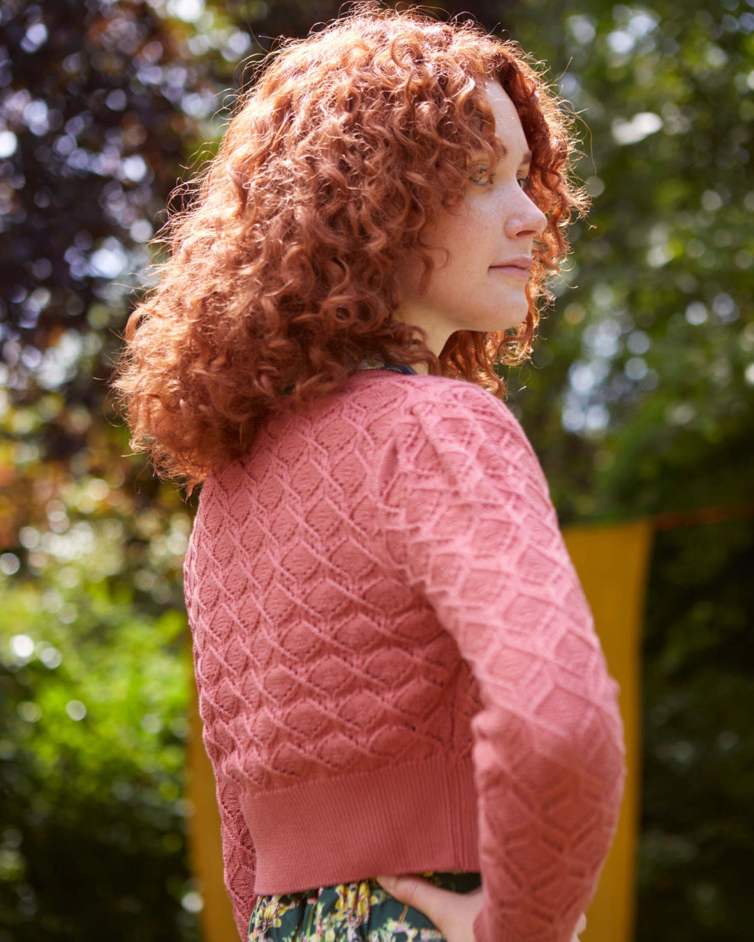 Ginger haired woman wearing a pink textured cardigan in a garden.