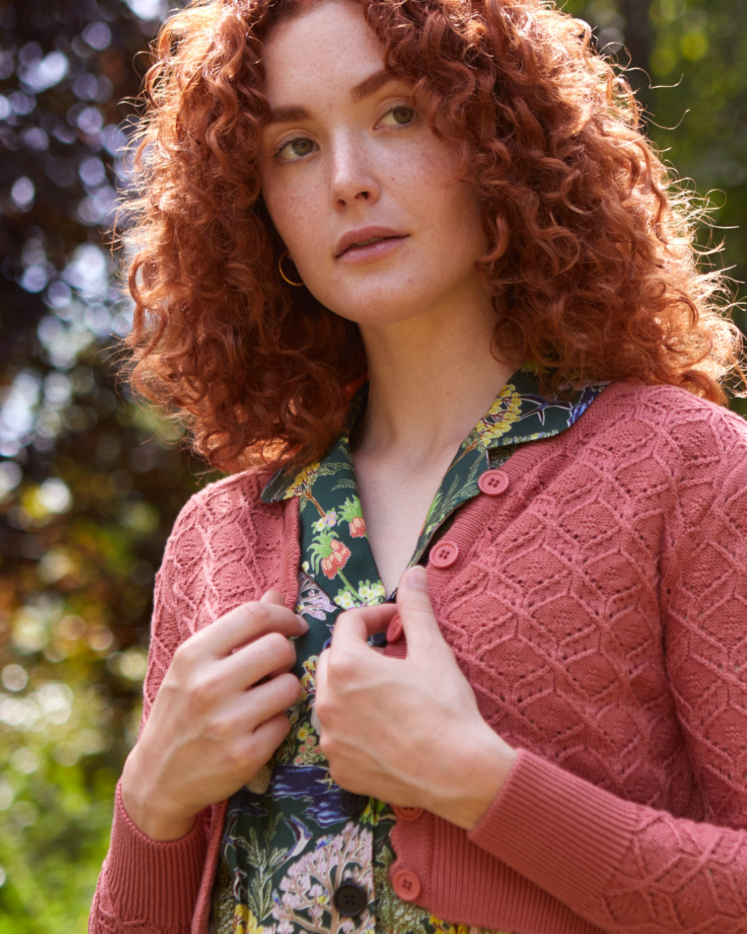 Ginger haired woman wearing a patterned, floral jumpsuit and a pink textured cardigan in a garden.