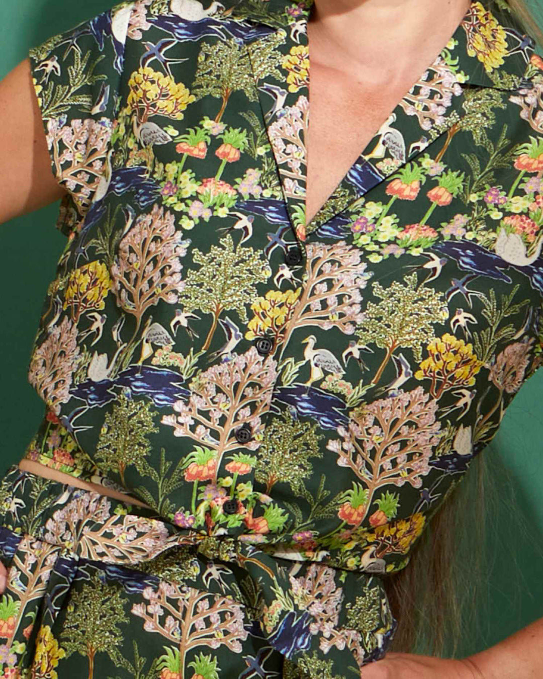 Close up of a front-tie top. The print depicts a botanical garden with differently shaped trees and bushes and has birds flying amongst them. 