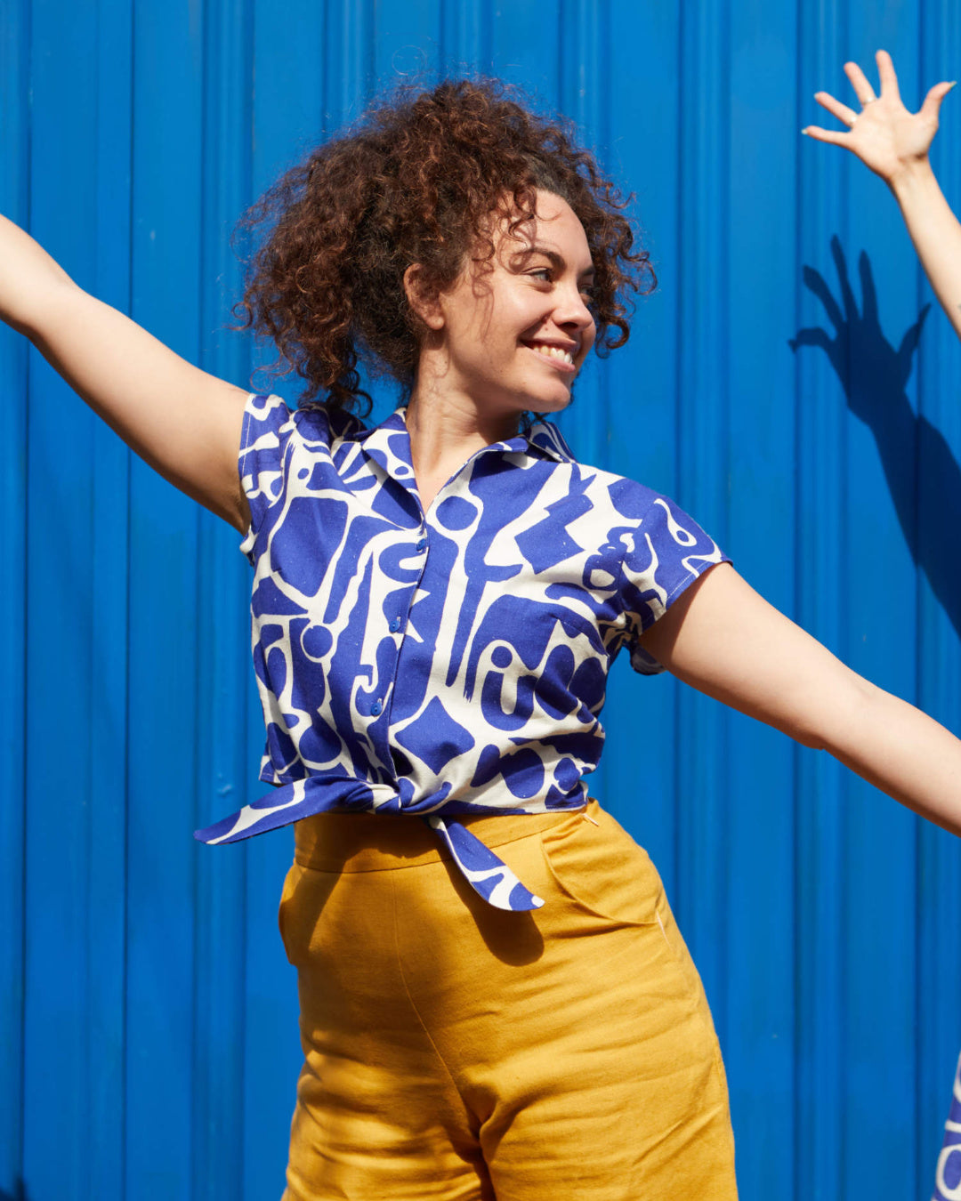 A woman stands in front of a blue background with her arms stretched out wide. She's wearing a blue and white, geometric printed front-tie top and yellow trousers.