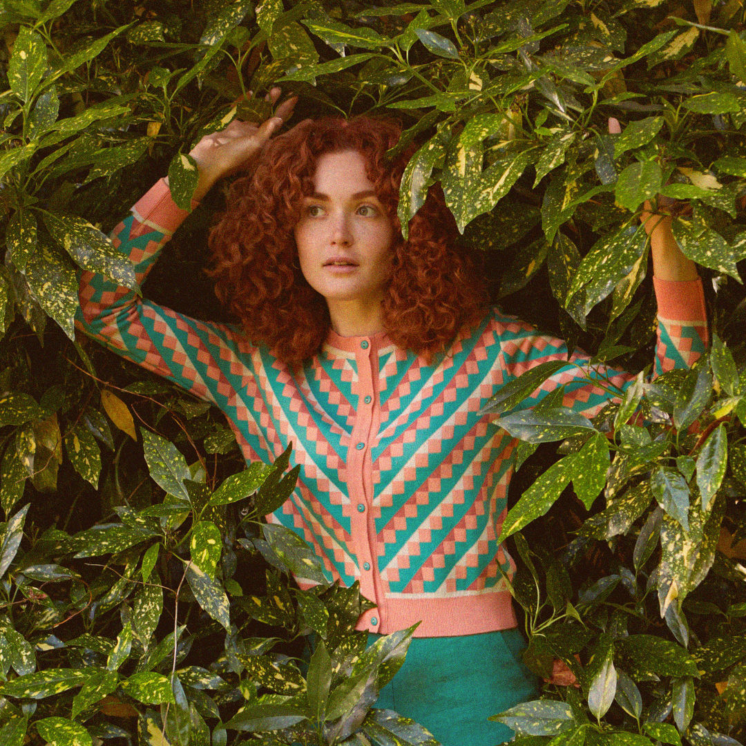 Woman coming out of the bushes, wearing a salmon and green checkered cardigan