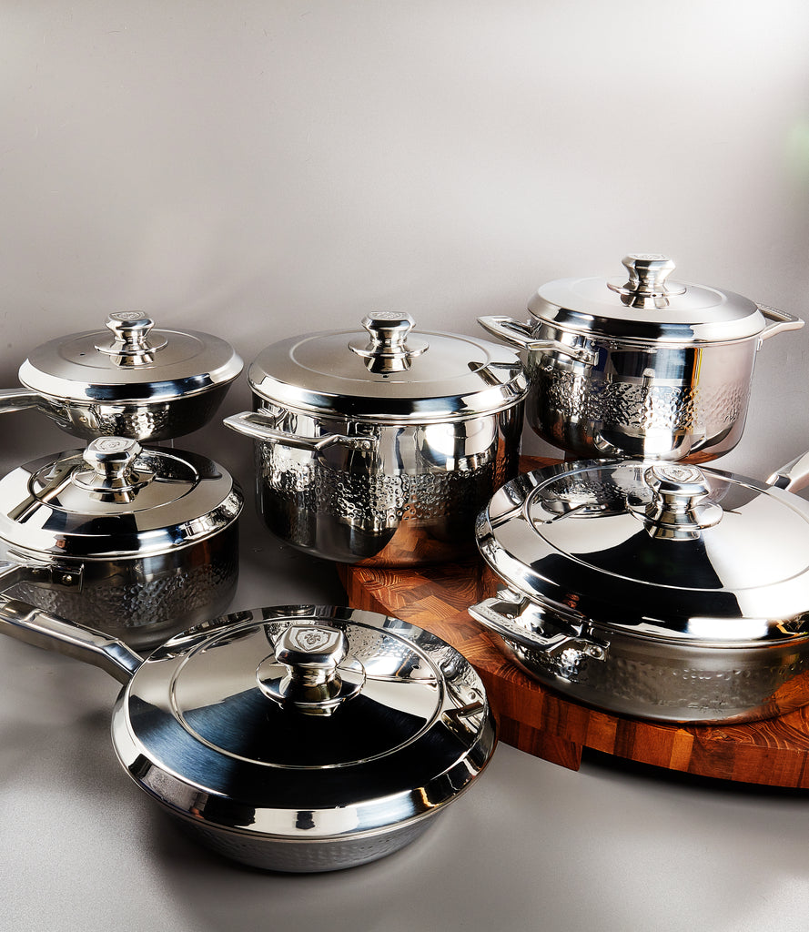 Stunning 12-pc collection of Dalstrong Cookware on a white set with a wooden cutting board underneath the three cookware.