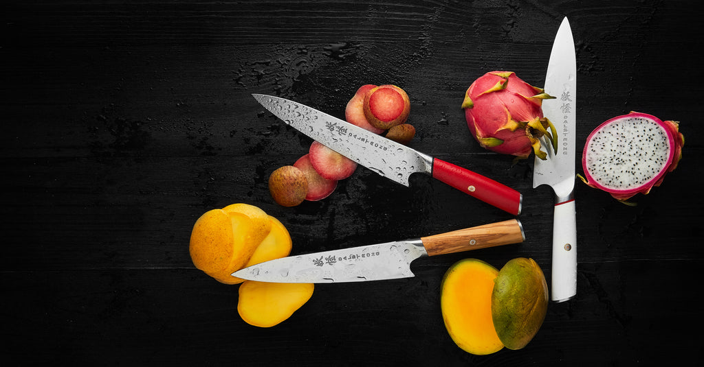 Dalstrong Phantom Series Chefs Knives in red, white, and olive wood handles on a black wooden table with slices of different fruits.