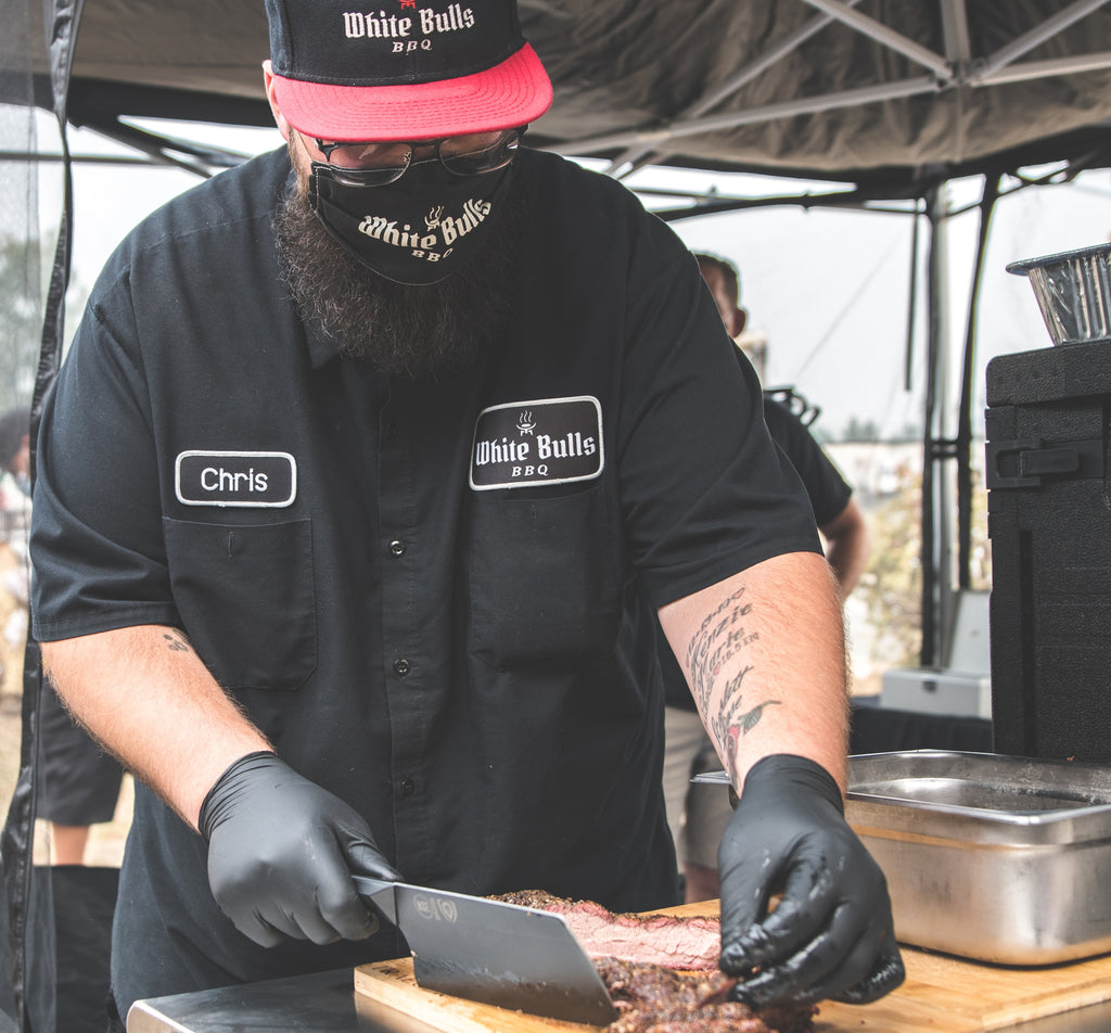 Chris Apple (@whitebullsbbq) using a black cleaver on a slab of meat outdoors 