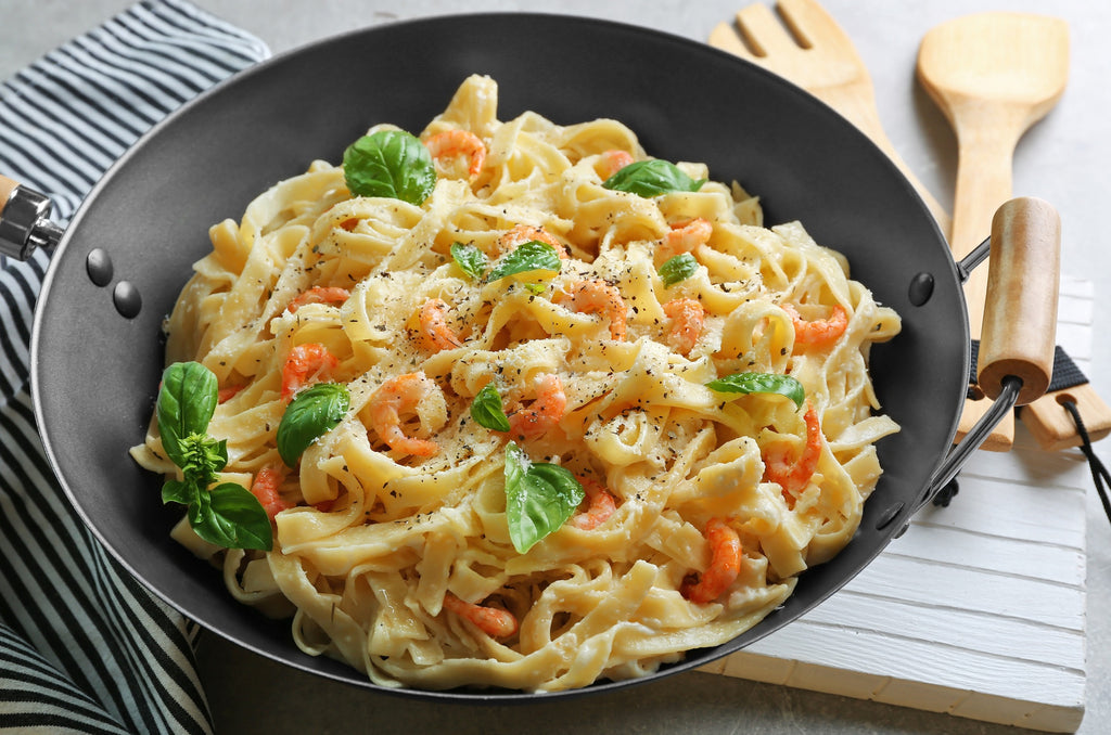 A dark grey wok filled with pasta covered in alfredo sauce made with parmesan cheese and colorful vegetables
