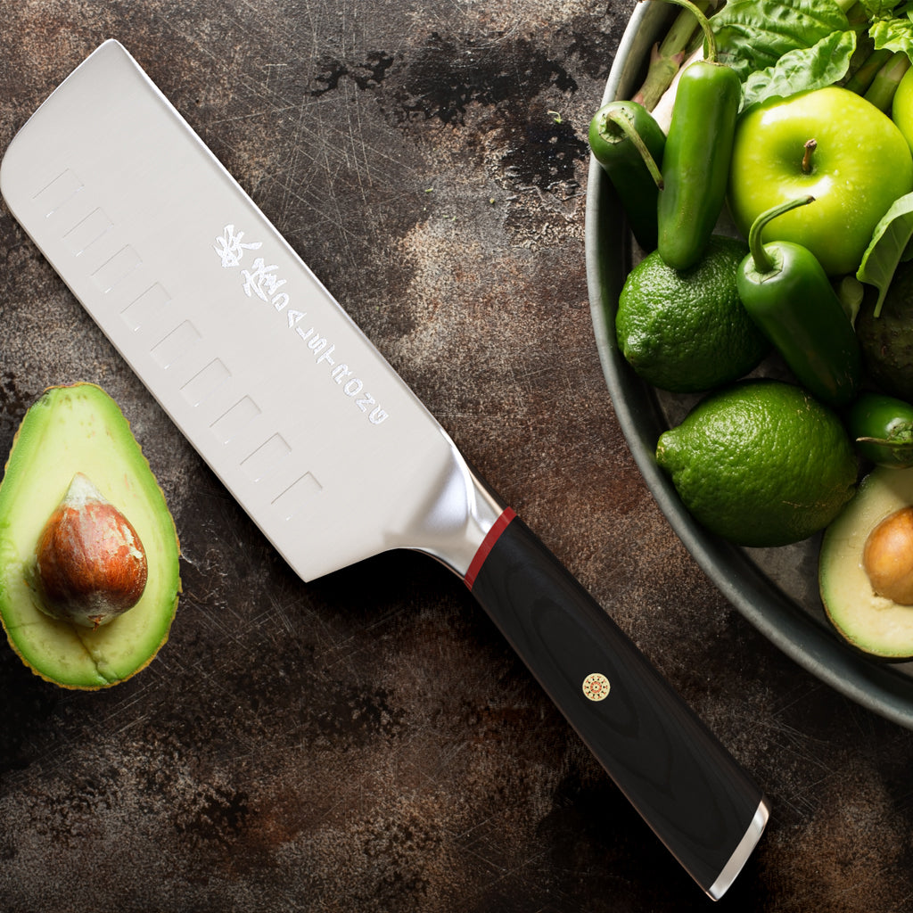 An avocado cut in half with the seed still intact next to a nakiri knife and bowl of green vegetables