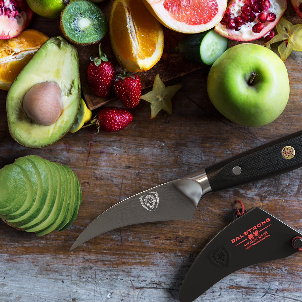 A selection of fruit on a dark surface next to a peeling knife and sliced avocado