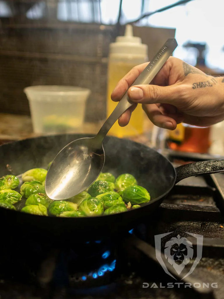 A man holding the Dalstrong © Professional Chef Tasting & Plating Spoon with cooked brussel sprouts on a pan.