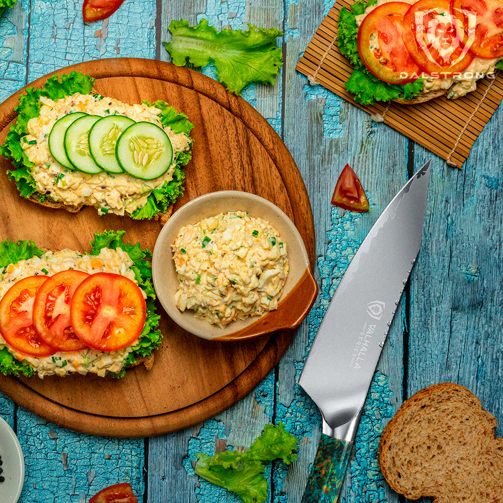 Egg Salad being assembled into a sandwich with lettuce, cucumber, and tomato on a wooden roun cutting board and a Dalstrong Valhalla Chef Knife on the side