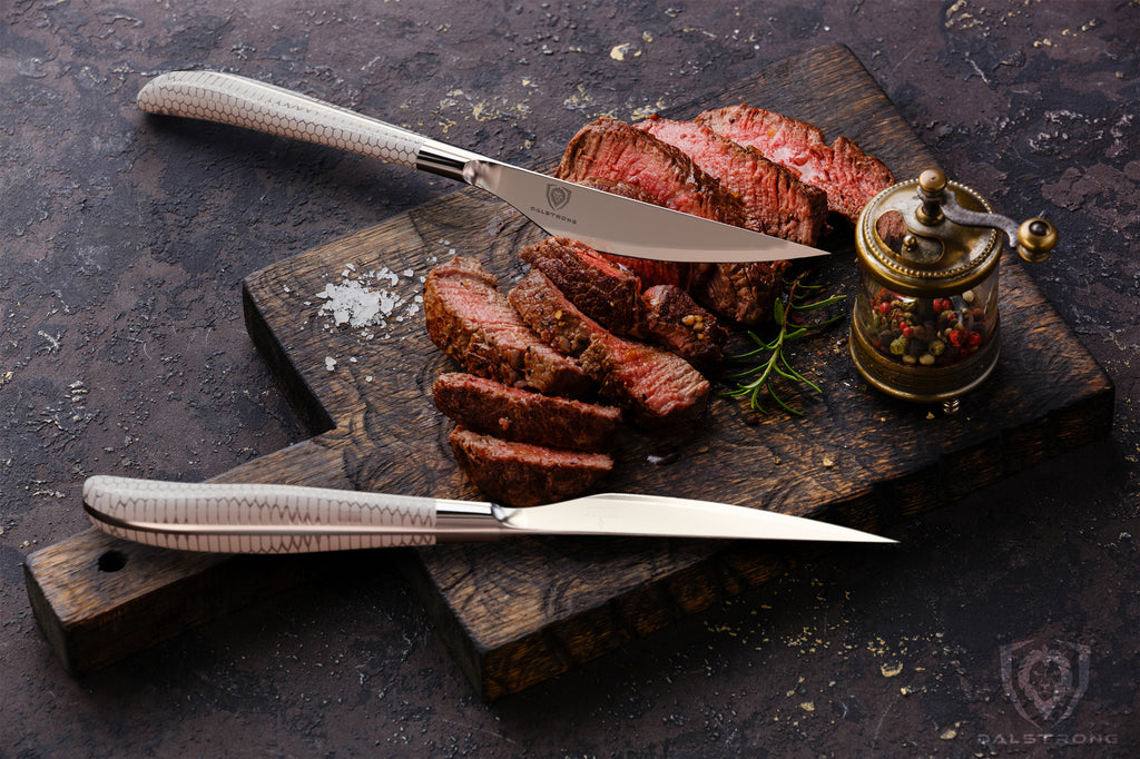 Two steak knives with white handles on a wooden cutting board next to chopped steak with a red center 