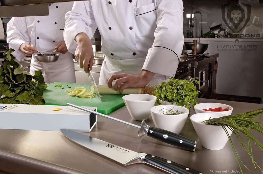 A professional chef chops food in the background while a honing steel rests on a box in the foreground 