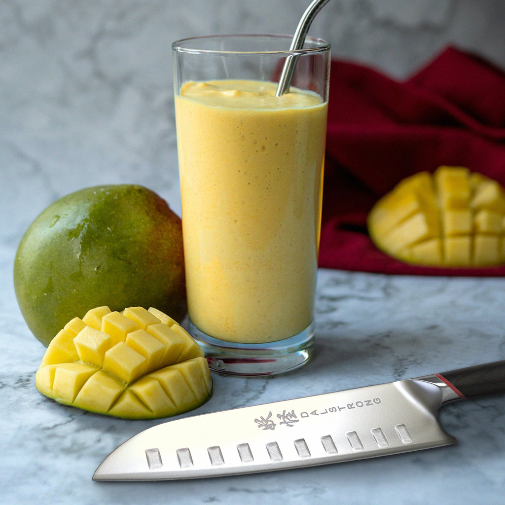 Chopped mango next to a whole mango and a glass of mango juice beside a santoku knife