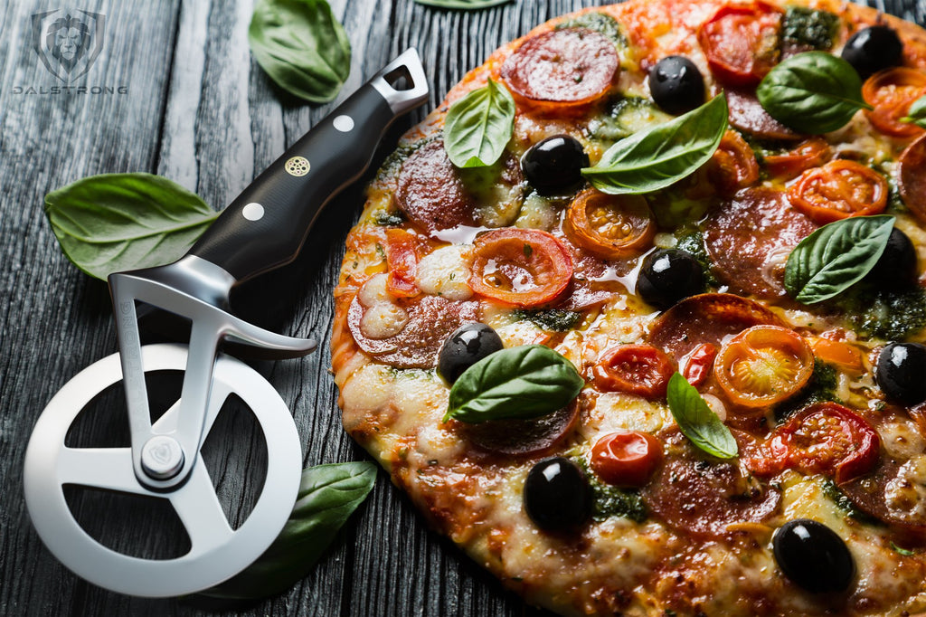 A pizza cutter on a wooden table rests beside a full cooked pizza that has olives tomatoes and basil as toppings