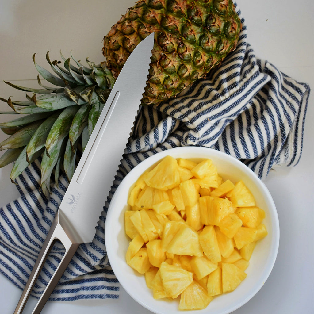 A stainless steel kitchen knife on a back and white kitchen towel next to a small bowl of chopped pineapple cubes