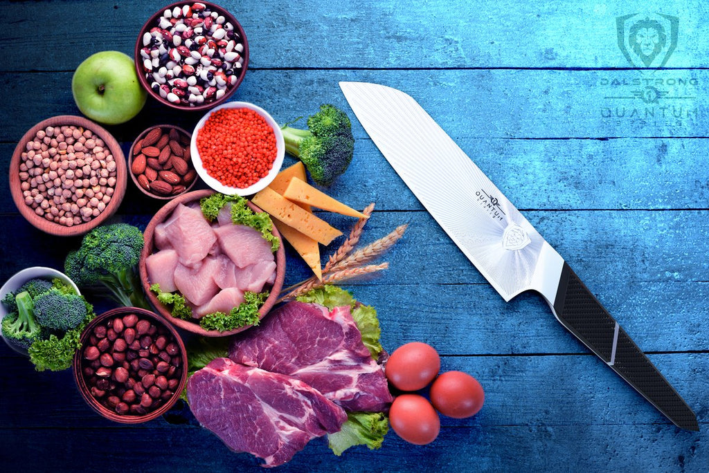 Quantum 1 Series santoku knife on a blue table next to a variety of food including fruit, vegetables and chicken